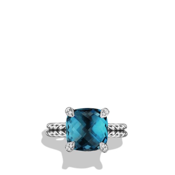 A silver ring with a twisted band featuring a large, square-cut blue gemstone in the center, secured by four prongs with small diamonds on each prong. Set against a plain white background, the ring casts a subtle shadow below it.