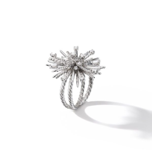 A silver ring with a twisted band features a unique burst design at the top, resembling an explosion of silver sticks adorned with small jewels. The ring casts a subtle shadow on a white background.