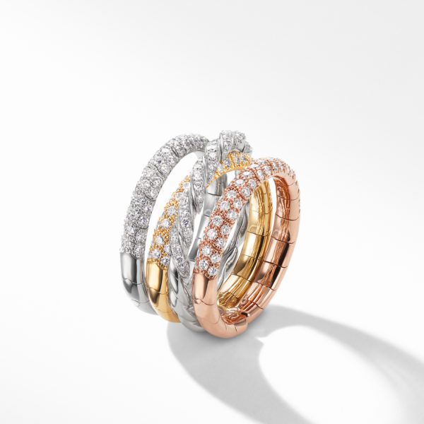 A stack of three intertwined rings made of white, yellow, and rose gold, each encrusted with diamonds, displayed on a reflective, light grey surface. The rings are elegantly arranged, showcasing their sparkle and intricate detailing.