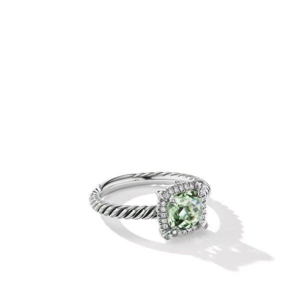 A close-up of a silver ring featuring a twisted band design and a large, rectangular green gemstone surrounded by smaller white gemstones in a square setting. The ring casts a soft shadow on a white surface.