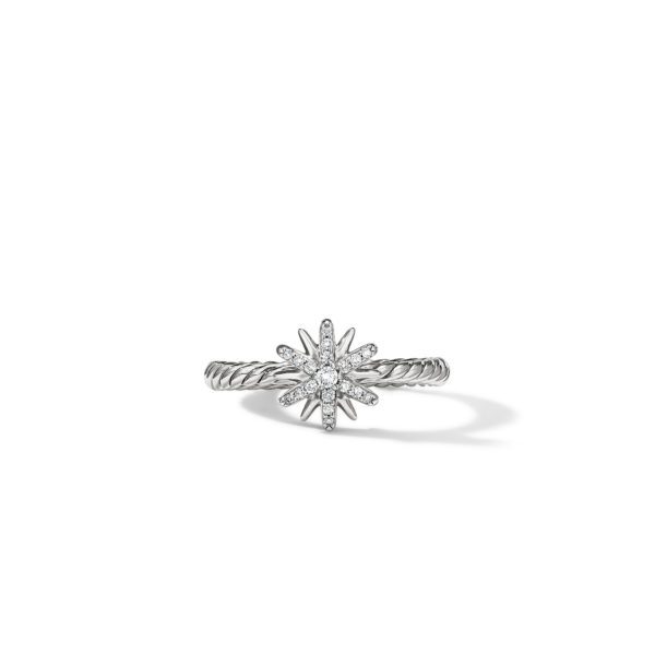 A silver ring with a starburst design as the centerpiece, encrusted with small, sparkling gemstones. The band has a braided texture. The ring casts a subtle shadow on the white surface underneath.