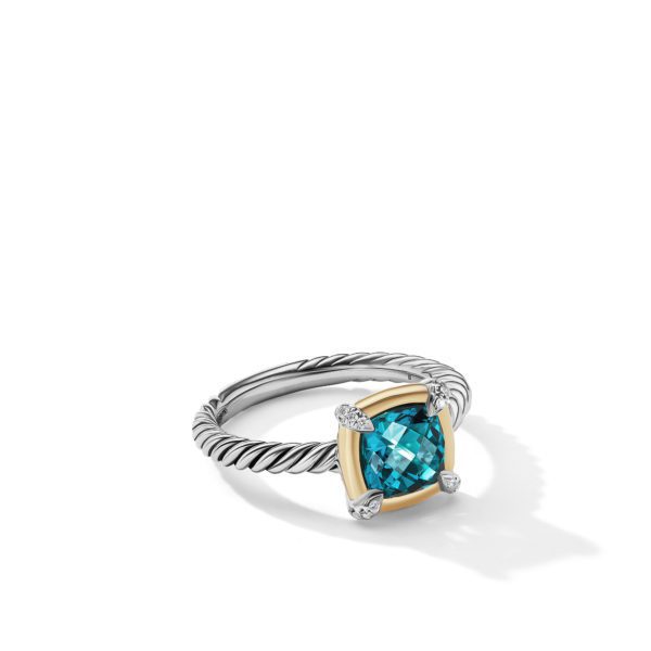 A silver ring with a twisted band is set with a square-cut blue gemstone in a gold bezel, further adorned with small diamonds at each corner. The ring casts a soft shadow on a white surface.