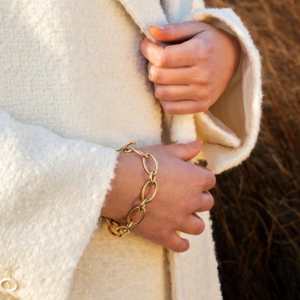 Close-up of a person wearing a white textured coat, with their hands clasped in front, showcasing the Roberto Coin Alternating Link Bracelet on the right wrist. The background appears to be a natural outdoor setting with earthy tones.