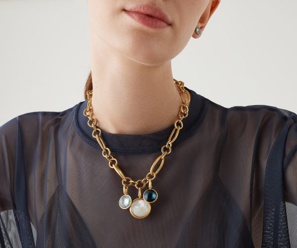 A person adorned in a sheer black top and sporting the Marco Bicego Jaipur Gold Mixed Link Necklace, which features three large round pendants including a pearl, poses against a neutral background. The person also accessorizes with a small earring.