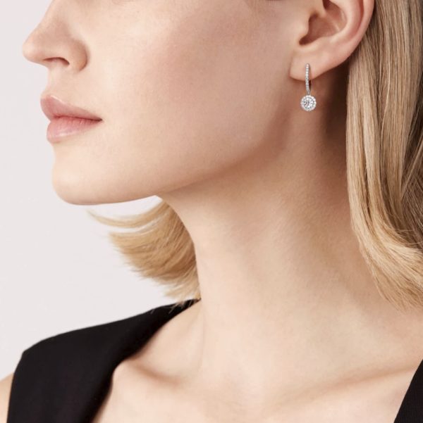 A close-up of a person's left profile shows their neck and partial face. They are wearing a black sleeveless top and a single Forevermark Center of My Universe Pavé Huggies with Halo Diamond Drops earring, featuring a hoop encrusted with small stones and a larger, round, stone-encrusted pendant. They have short, blonde hair.