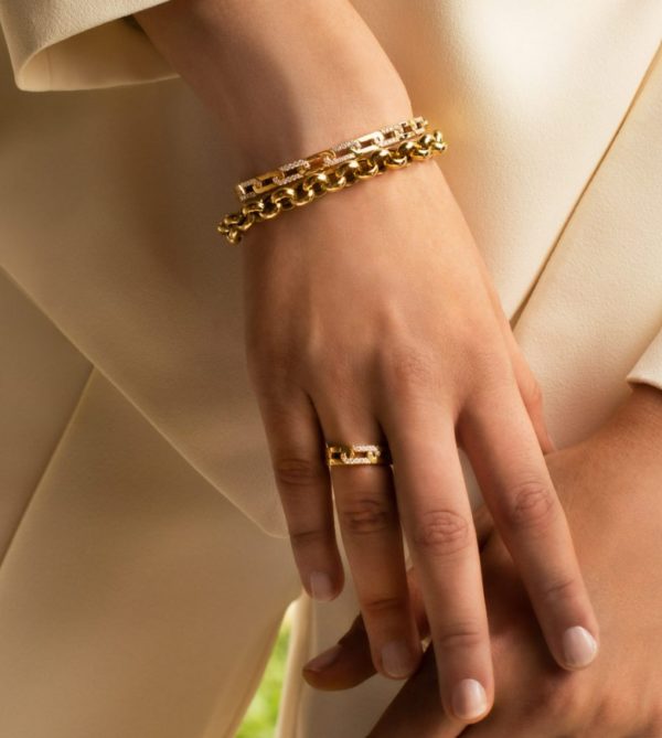 Close-up of a person's hand adorned with the Roberto Coin Navarra Diamond and Gold Link Ring, accompanied by a matching gold chain bracelet. The individual is dressed in light-colored, elegant clothing. The softly blurred background accentuates the stunning jewelry.