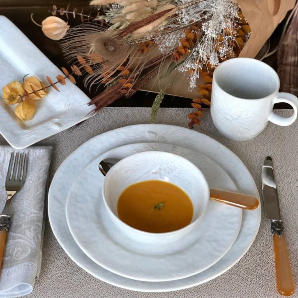 A beautifully set table features a white bowl of orange soup on a Carmel Ceramica Foresta Dinner Plate, accompanied by a fork and knife with wooden handles. A neatly folded napkin rests to the side, and a white mug is placed nearby. Dried flowers serve as a decorative centerpiece.