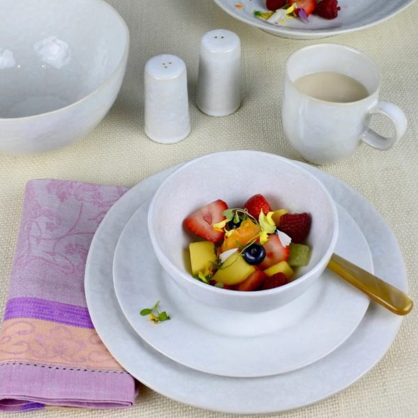 A white bowl filled with a colorful fruit salad, including strawberries, blueberries, and pineapple, is set on a Carmel Ceramica Cozina Dinner Plate - White. Nearby, a cup of milk, salt and pepper shakers, and a folded lavender napkin lie on a textured beige tablecloth.