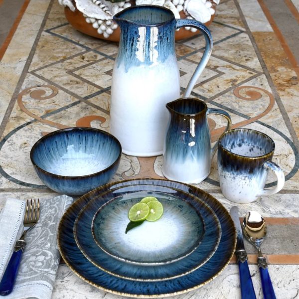 A set of gradient blue and white ceramic dishes arranged on a patterned stone table, featuring the Carmel Ceramica Cypress Grove Salad Plate. The collection includes a large pitcher, a small jug, a bowl, a mug, and three nested plates with lime slices on the top plate. The table is also adorned with blue-handled cutlery, linen napkins, and a decorative element in the background.