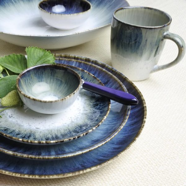 A set of blue and white gradient ceramic dishes, including a Carmel Ceramica Cypress Grove Mug, plates of various sizes, and small bowls arranged on a light woven surface. A small purple-handled butter knife, a green leaf, and a strawberry are placed on the largest plate.