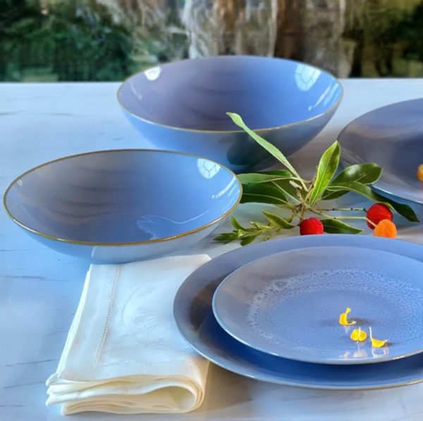 A table setting featuring the Carmel Ceramica Rhapsody Blue Bowl - Medium and matching blue plates with gold rims. A white cloth napkin is folded next to the dishes, and a sprig of leaves with small red and orange berries adorns the scene. The background shows an outdoor view with greenery.