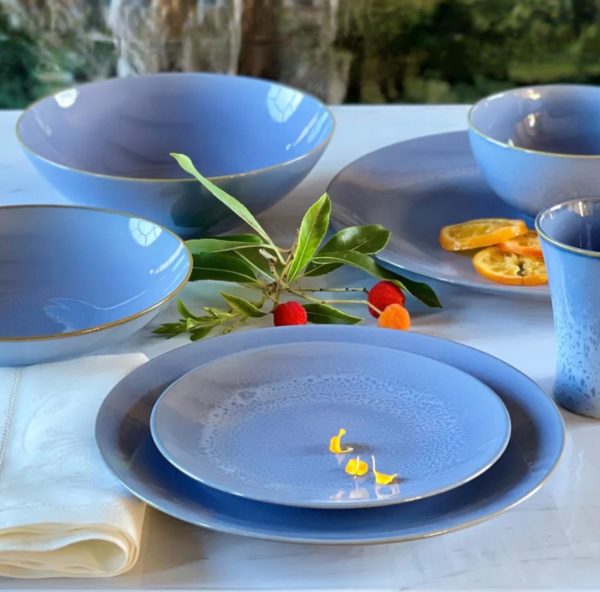 The Carmel Ceramica Rhapsody Blue Salad Plate, displayed on a white surface as part of a cohesive dinnerware set that includes plates, bowls, and a cup. The arrangement is enhanced by green leaves, red and orange berries, and orange slices, with a crisp white napkin elegantly placed under one of the plates. A blurred outdoor background adds to the serene setting.