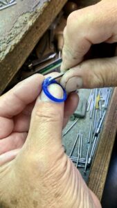 A pair of hands works on shaping a blue ring with a tool at a jewelry workbench. Various metal rods and tools are scattered on the workbench surface in the background.