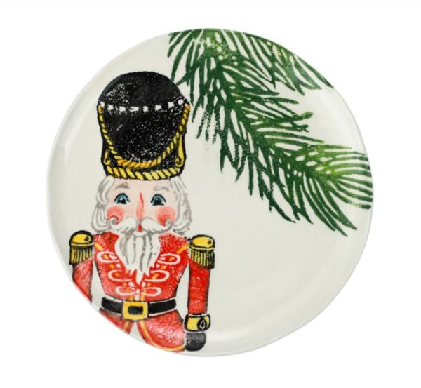 The Vietri Nutcrackers Red Salad Plate is a round dish adorned with a festive design showcasing a Nutcracker soldier in a red uniform with gold details, featuring a white beard and black hat. In the upper right corner, there’s a green pine branch, set against the plate's white background.