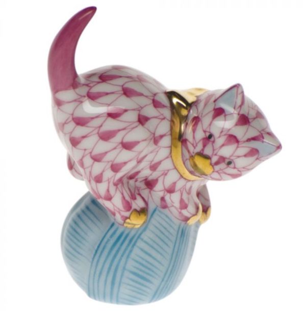 Presenting the Herend Mischievous Cat Figurine in Raspberry: a porcelain masterpiece featuring a pink and white patterned cat with elegant gold accents. The playful feline is whimsically poised atop a light blue ball of yarn, with its tail raised high and front paws clutching the yarn.