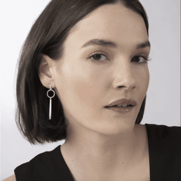 A person with short, dark hair is wearing a black top and a pair of LAGOS Caviar Spark Beaded Circle Diamond Drop Earrings. The background is plain white. The person has a faint smile and is looking directly at the camera.