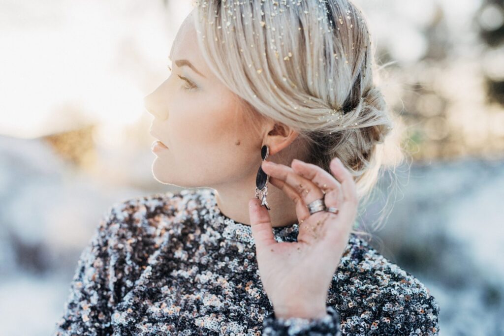 A beautiful woman in a sweater with snow in her hair showing off a pair of earrings and rings