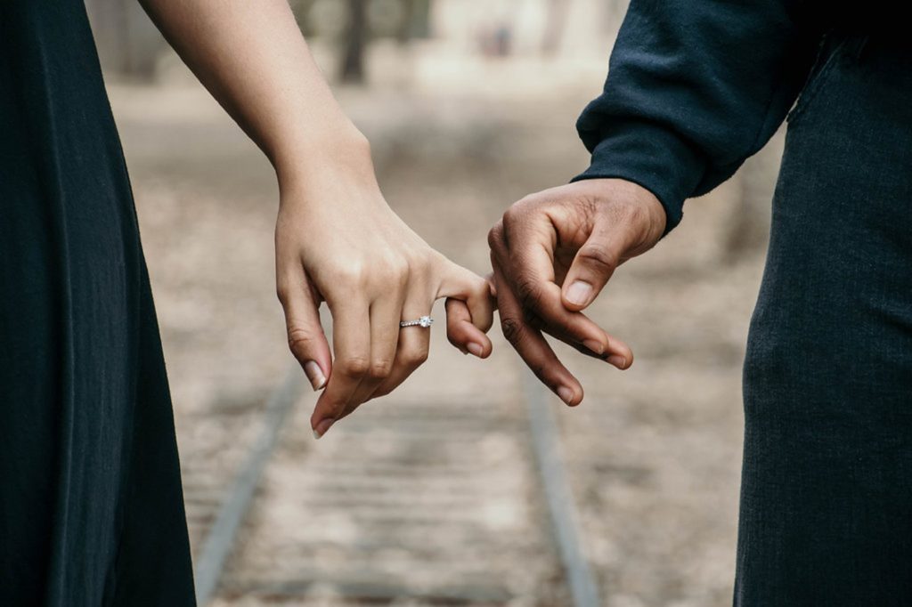 An engagement photo of a couple’s hands linked at the pinky with train tracks in the background.