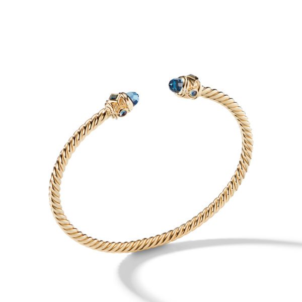 A twisted gold rope bracelet with open ends capped by blue gemstone accents, each set in a detailed gold setting. The bracelet is suspended slightly above a white surface, casting a soft shadow.