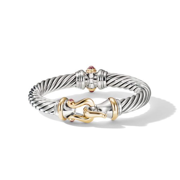 A twisted silver bangle bracelet featuring gold accents and adorned with small red gemstones. The bracelet has an ornate clasp and a combination of smooth and textured elements. It casts a subtle shadow on the white background.