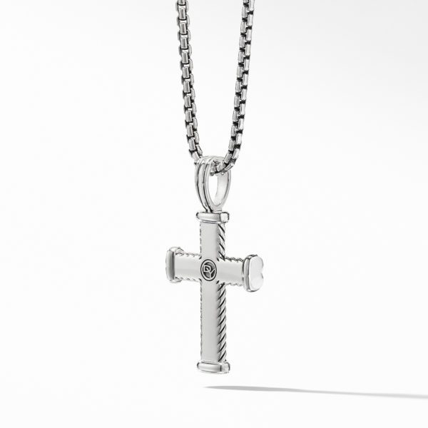 A close-up of a silver cross pendant hanging on a silver chain against a light gray background. The cross has a sleek design with subtle ridges along the edges and a small, circular emblem in the center.