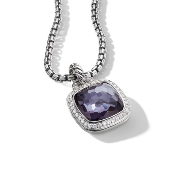 A silver necklace features a cushion-cut purple gemstone pendant, bordered with small, clear crystals. The pendant hangs from a rounded box chain, and both pendant and chain are displayed against a white background, casting a subtle shadow.