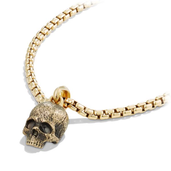 A gold chain necklace featuring a detailed skull pendant. The skull has a textured, antique finish, and the chain is composed of small, interlocking rectangular links. The pendant hangs from a rounded gold loop attached to the chain.