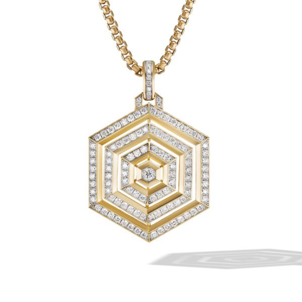 A gold pendant necklace featuring a hexagonal, three-dimensional design adorned with rows of small, sparkling diamonds. The pendant's intricate pattern forms concentric shapes, hanging from a gold chain. A shadow of the pendant is visible underneath.