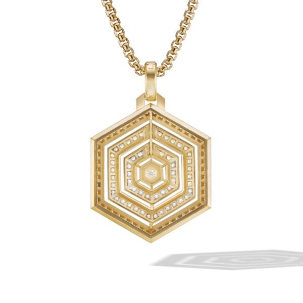 A gold necklace with a hexagon-shaped pendant featuring intricate geometric patterns and small, embedded gemstones. The pendant casts a subtle shadow to the lower right, highlighting its detailed design.