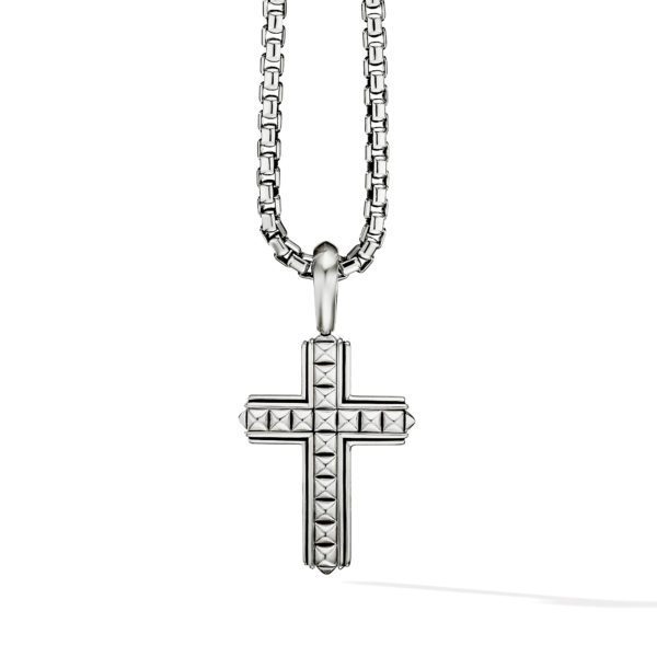 A silver cross pendant with a geometric pattern hangs from a silver chain. The cross features small, pyramid-shaped studs and a polished finish. The chain is made up of square-shaped links. Both the pendant and chain shine under the light, highlighting their intricate design.