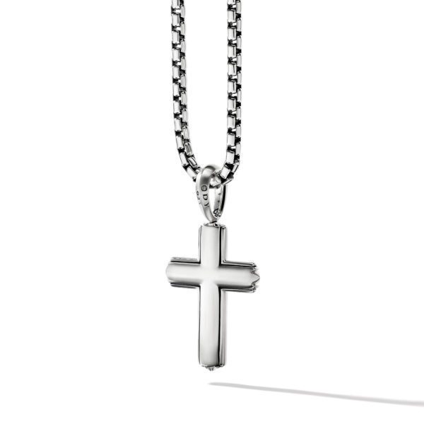 A silver cross pendant hangs on a silver chain against a white background. The cross has a simple, sleek design with smooth edges. The chain has a box link pattern, and the pendant casts a shadow on the surface below.