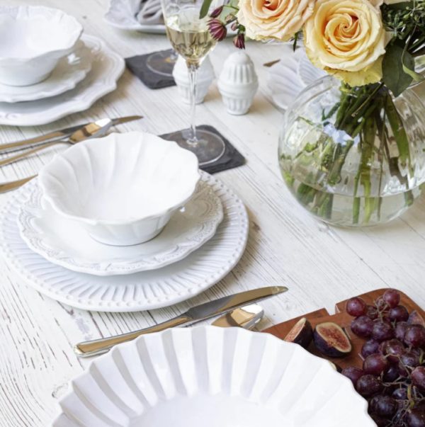 A white, rustic dining table is set with elegant white dishware, including the Vietri Incanto Ruffle Cereal Bowl - White for added charm. Two champagne glasses and a vase with yellow roses contribute to the sophisticated atmosphere. A wooden board with grapes and figs sits on the side, enhancing the inviting ambiance.