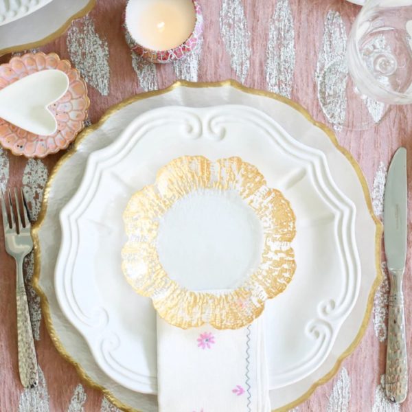 The table setting features a Vietri Incanto Baroque Dinner Plate in white, topped with a smaller white plate adorned with a gold flower-shaped border. Beneath the small plate lies a white cloth napkin. A knife is placed to the right and a fork to the left of the plates. In the background, there is a lit candle and various decorative items.