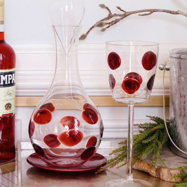 A Vietri Drop Wine Glass - Red, decorated with large red polka dots, sits on a white tabletop beside a matching glass decanter. Positioned next to them is a bottle of Campari and a silver ice bucket. Nearby, some green foliage adds a touch of nature, while a branch with small twigs hangs above in the background.