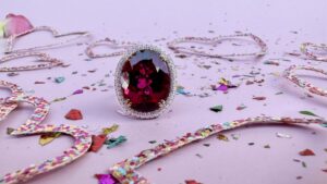 A large, round-cut red gemstone ring adorned with a halo of smaller diamonds is placed on a light pink surface. Colorful confetti and sequins are scattered around, along with decorative strands of sequins, adding a festive touch to the scene.