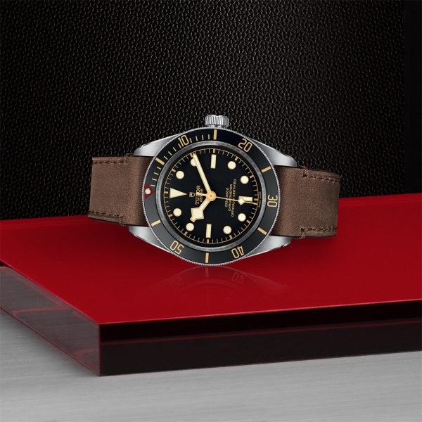 The TUDOR Black Bay Fifty-Eight, a luxury watch with a black dial, cream indices, and brown leather strap, is showcased on a red surface against a textured black background. It features golden hour markers and a rotating bezel.