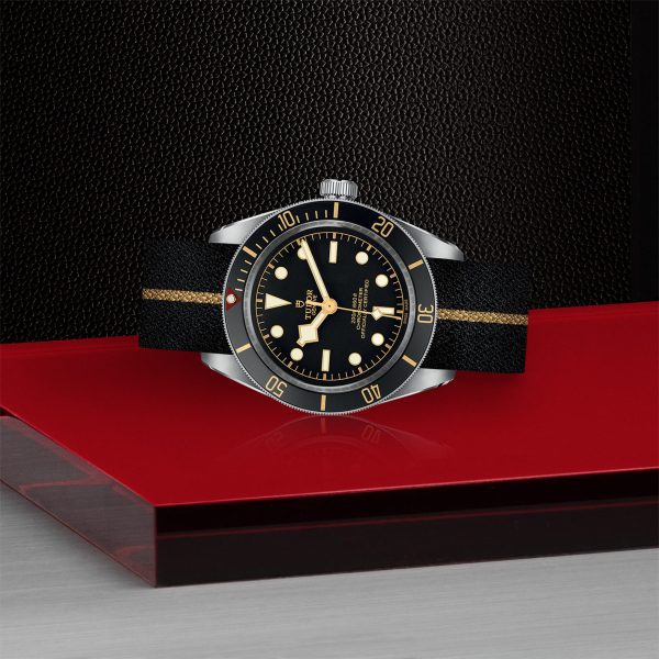 The TUDOR Black Bay Fifty-Eight, featuring a sophisticated black and gold dial, is showcased on a striking red platform against a textured black background. The watch boasts a sleek black strap with an elegant gold stripe in the center and gold hour markers. The hands and bezel are adorned with refined gold accents.