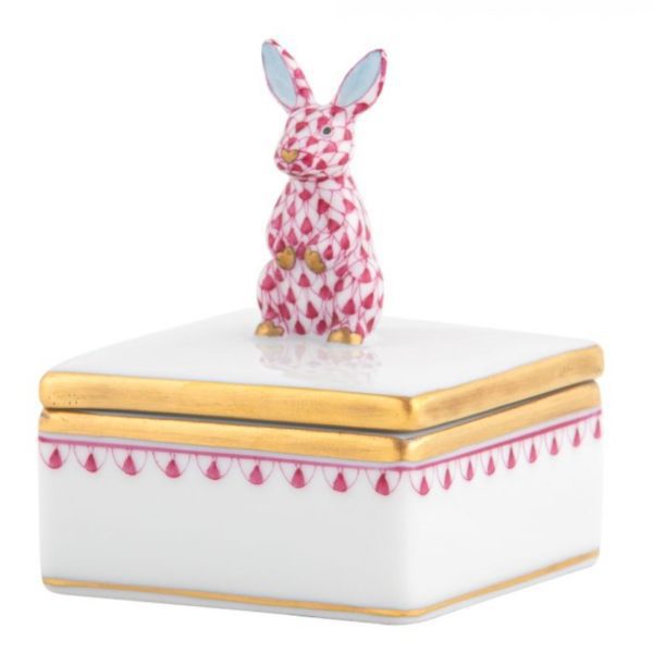 The Herend Bunny Box Figurine - Raspberry is a white, square trinket box adorned with a delicate gold trim along the lid and edges. The lid features a small, checkered-patterned pink rabbit figurine sitting on top. The sides of the box are decorated with pink and gold scallop designs.