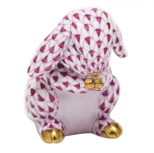 The Herend Praying Bunny Figurine - Raspberry is a porcelain figure of a rabbit with its head bowed. It features a detailed, patterned design in alternating shades of white and raspberry with gold accents on its feet and yellow on its chest. The body is adorned with fish scale-like patterns.