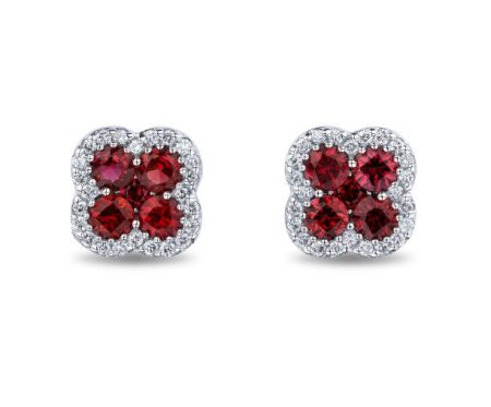 A pair of stud earrings featuring a flower-shaped design with four round ruby gemstones in the center, surrounded by small clear diamonds. The metal setting appears to be silver or white gold. The earrings are symmetrically positioned against a white background.