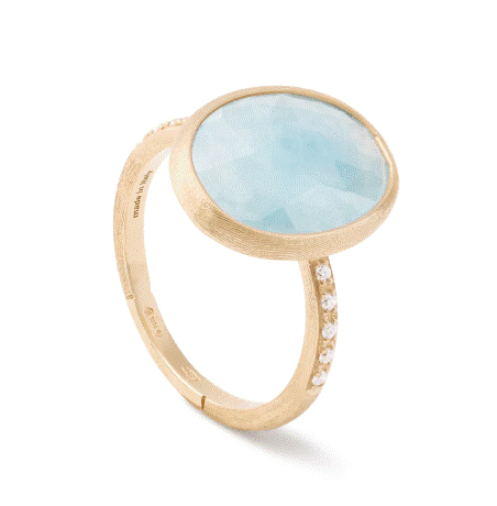 The Marco Bicego Siviglia Aquamarine and Diamond Ring showcases a large, circular light blue aquamarine gemstone at the top, complemented by a gold band adorned with small white diamonds along one side. The design is elegant and minimalist.