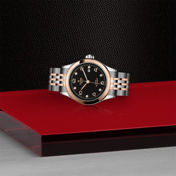 Silver and rose gold Tudor watch laying sideways on a red display platform