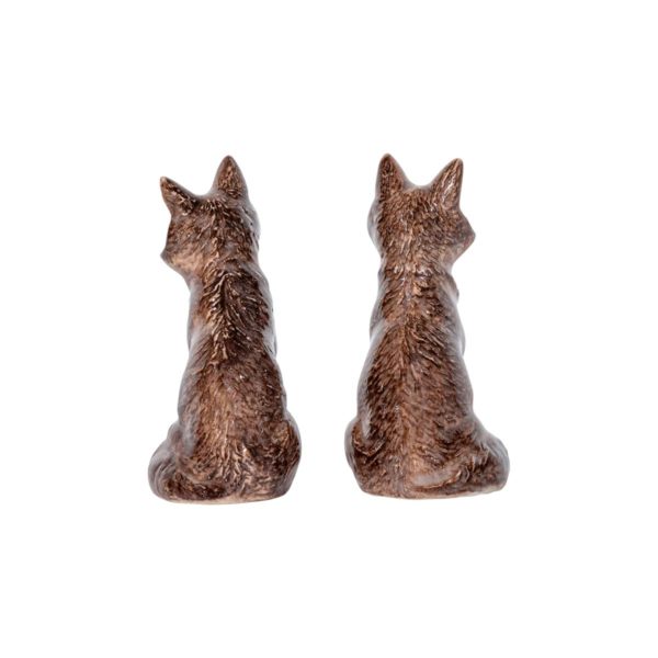 Two small, dark brown ceramic fox figurines from the Juliska Clever Creatures Fox Salt and Pepper Set sit side by side facing away, showcasing their detailed fur texture and pointed ears. The figurines have a realistic, rustic design and are positioned against a plain white background.