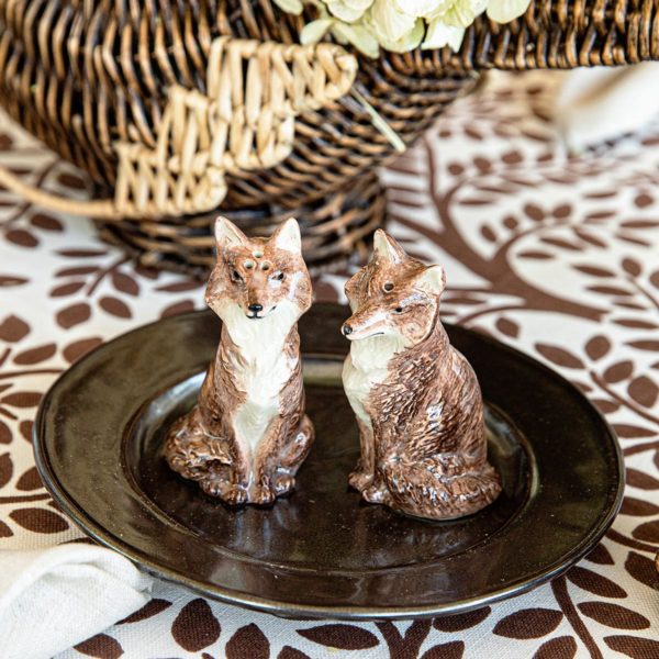 Two ceramic figurines from the Juliska Clever Creatures Fox Salt and Pepper Set, featuring brown and white foxes, sit on a dark-colored plate. The plate is placed on a tablecloth with a brown leafy pattern. In the background is a wicker basket filled with flowers, creating a rustic and charming aesthetic.