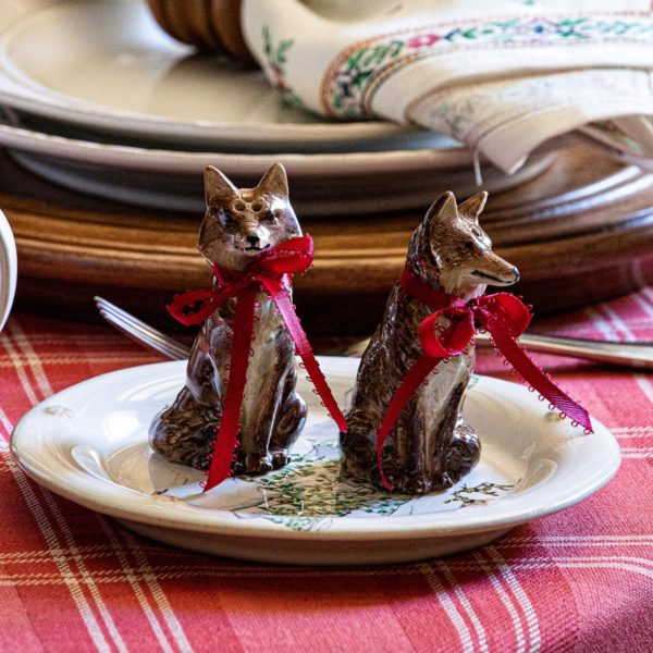 Two ceramic fox figurines with red ribbon bows around their necks, part of the Juliska Clever Creatures Fox Salt and Pepper Set, sit on a decorative plate. The plate is placed on a table set with a red checkered tablecloth, cutlery, and stacked plates in the background. The scene suggests a festive or holiday setting.