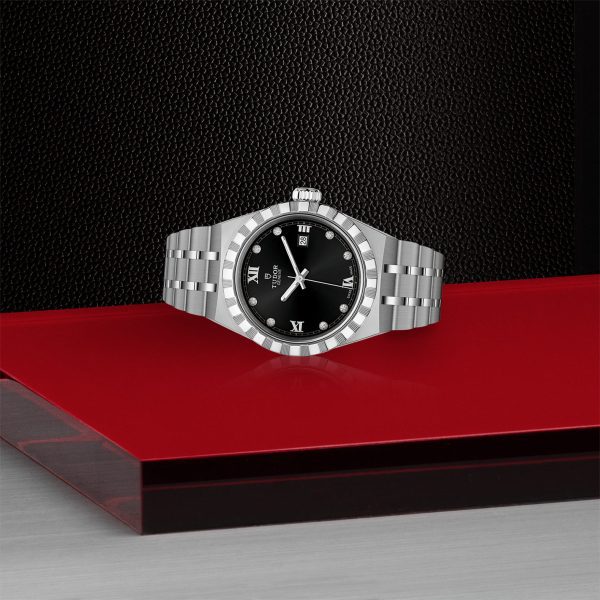 A TUDOR Royal silver wristwatch with a black dial showcasing a mix of Roman numerals and diamond markers is elegantly positioned on a red and black modern surface. The watch features a metal link band and a fluted bezel, exuding luxury design.