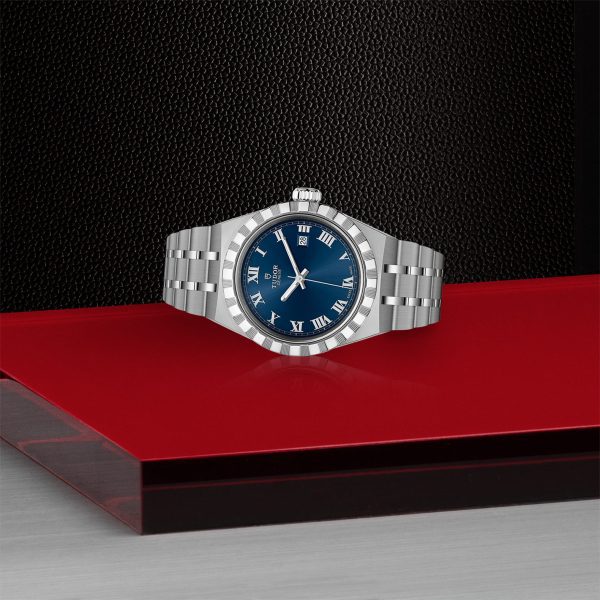 The TUDOR Royal is a luxury silver wristwatch featuring a blue dial, Roman numeral hour markers, and a date display at 3 o'clock. It is showcased on a red and black surface with a textured background.