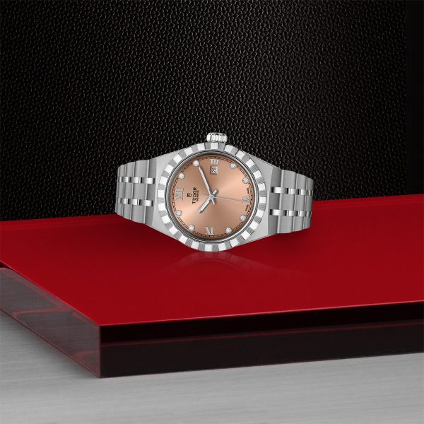 The TUDOR Royal, a luxury wristwatch with a stainless steel bracelet and a salmon-colored dial, is displayed on a red and black background. The watch features a date window and silver Roman numerals at the 12, 3, 6, and 9 o'clock positions.