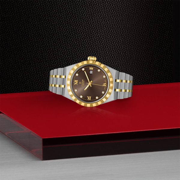 The TUDOR Royal, a luxury wristwatch with a gold and silver metal band, is displayed on a red and black surface. The watch features a gold bezel, a brown dial, and Roman numerals marking 6 and 12 o'clock, with a date window positioned at the 3 o'clock mark.