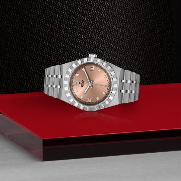 The TUDOR Royal, a luxury silver wristwatch featuring a metal link band and a round, light brown face, is displayed on a geometric surface adorned with black, red, and silver panels. The watch showcases Roman numerals and small crystal markers, seamlessly blending elegance with precision.